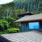 A house with gray shingles surrounded by lush green trees and mountains in the background.