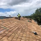 Workers installing shingles on a large roof under a partly cloudy sky.